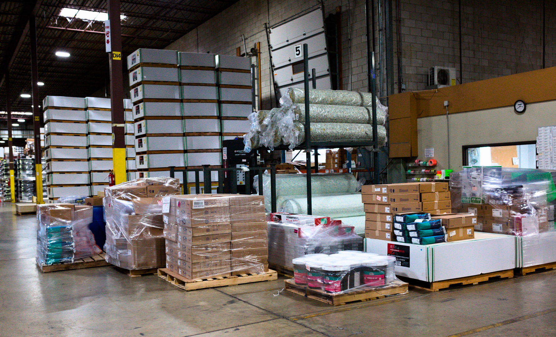 Warehouse area with stacked pallets, carpet rolls, and boxed goods.
