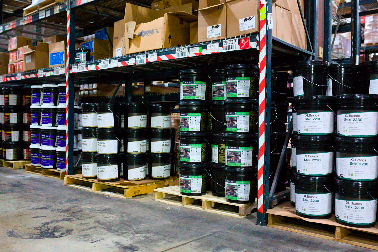 Warehouse shelves with stacked black adhesive buckets and cardboard boxes.
