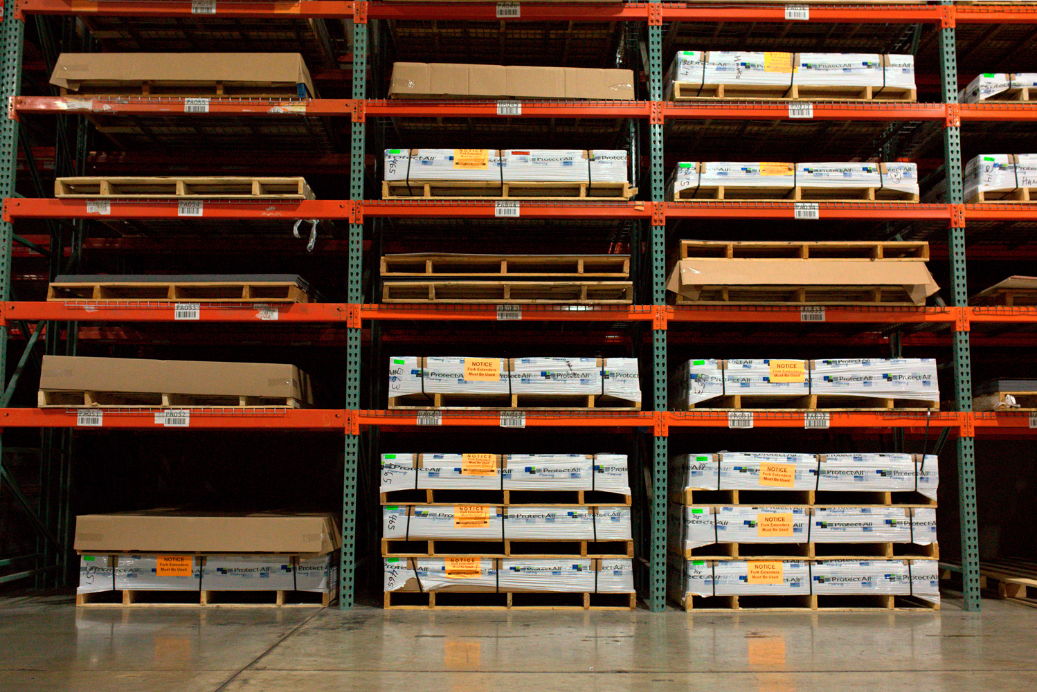 Warehouse racks with stacked Protect-All flooring pallets and cardboard sheets.