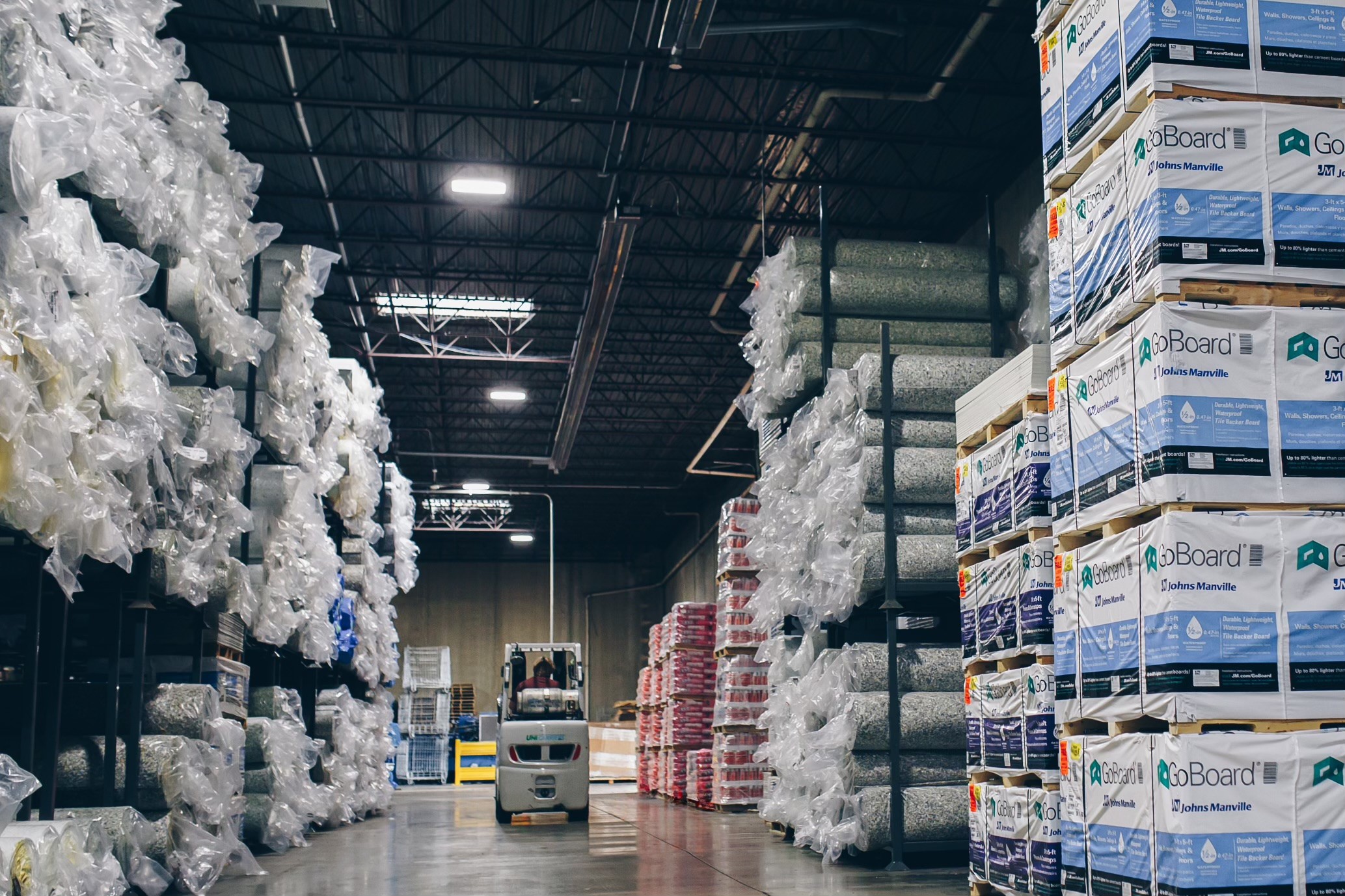Warehouse aisle with stacked flooring materials and a forklift in motion.